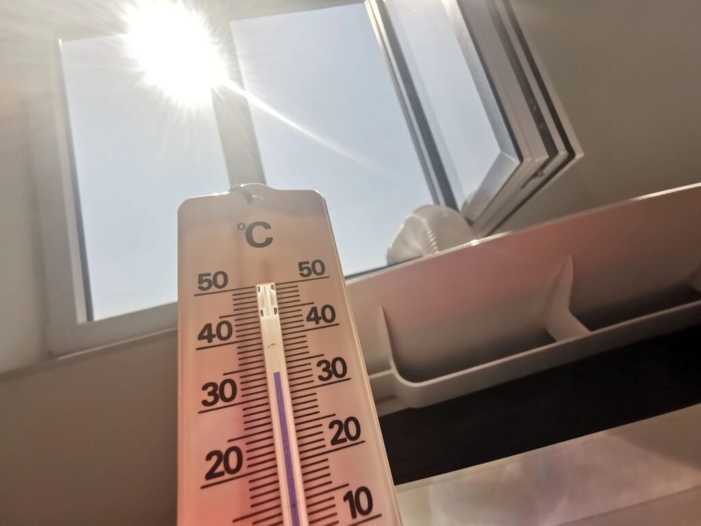 Analog celsius thermometer shows almost 35 degrees next to a mobile air conditioner on an open window with sun